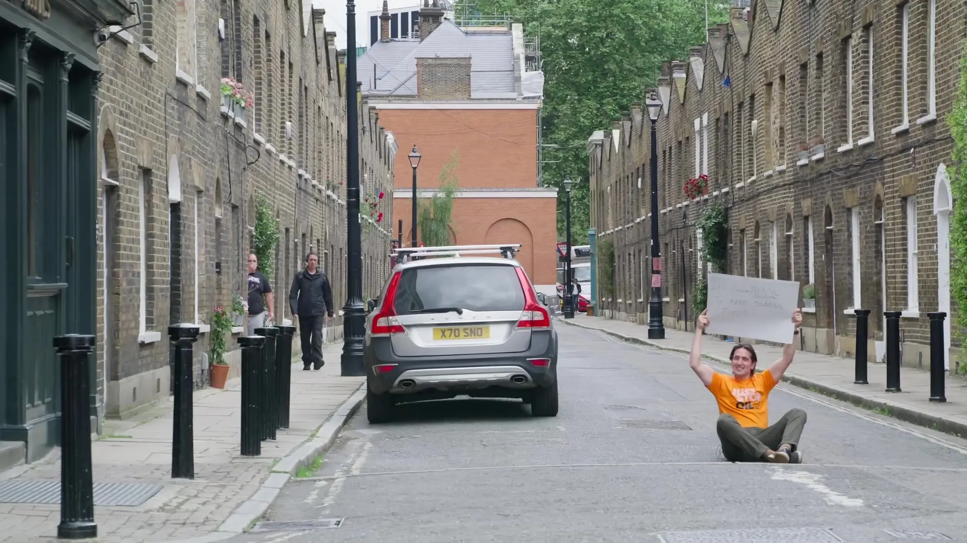 Graham sits in the middle of a quiet terraced street holding up his sign, a single car waiting behind him