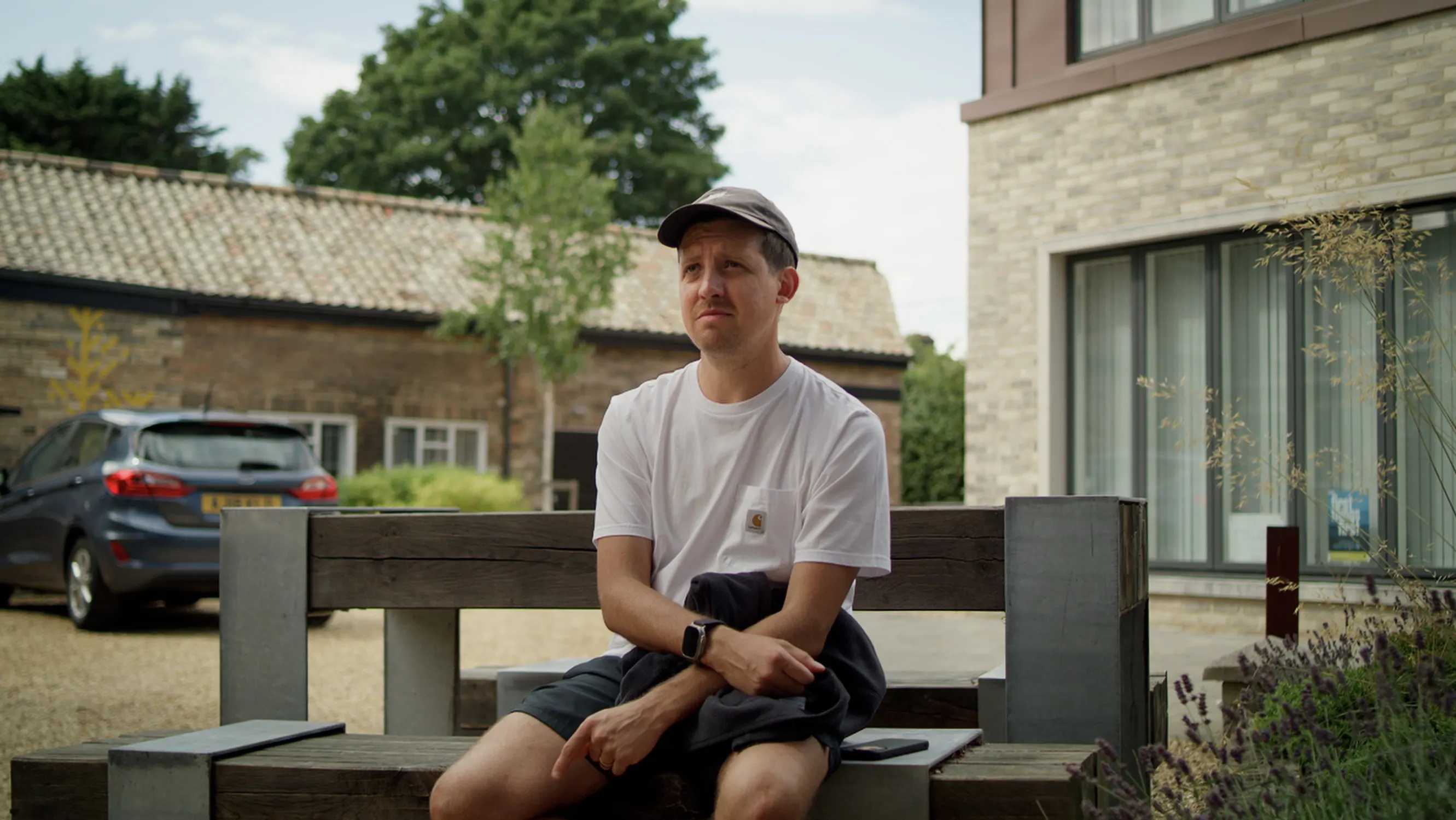 Mark G Smith as Peter, sitting on a bench outside a brick building, looking unconvinced