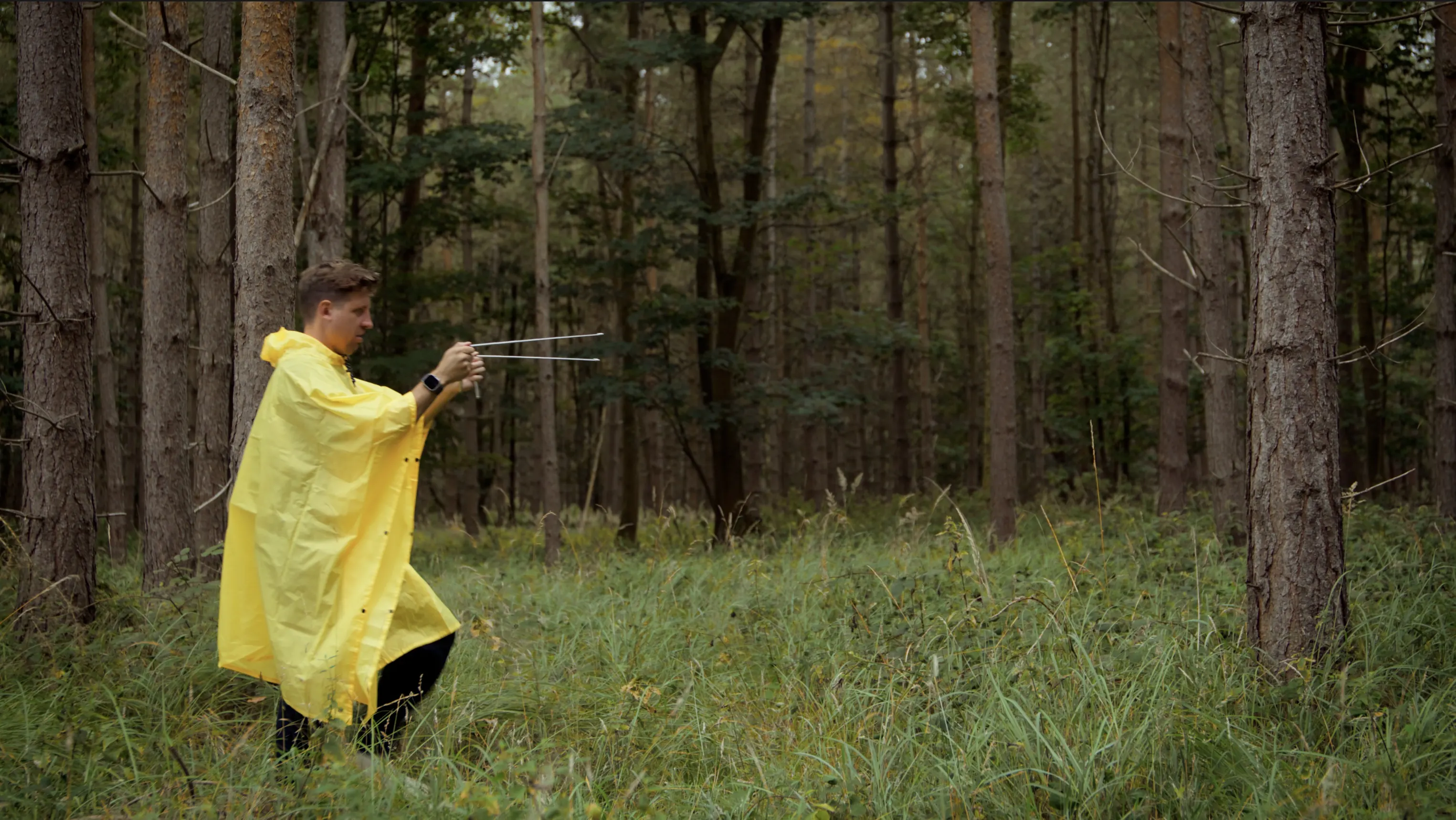 Mark G Smith as Peter, in a yellow poncho, walks through a pine forest holding dowsing rods