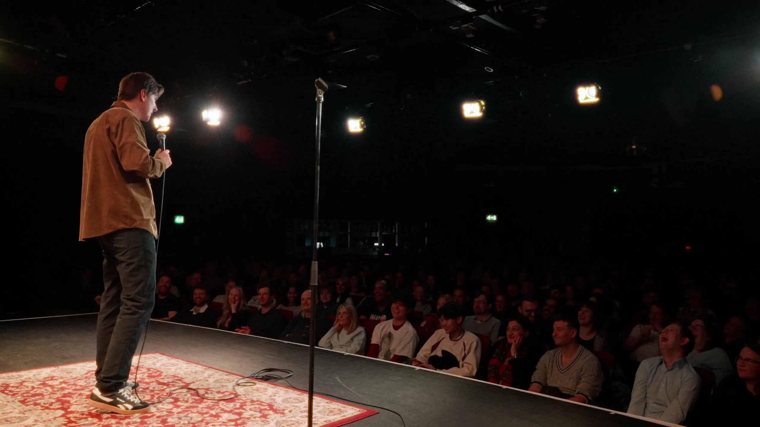 Sean McLoughlin on stage at Leicester Square Theatre, seen from behind towards a full house