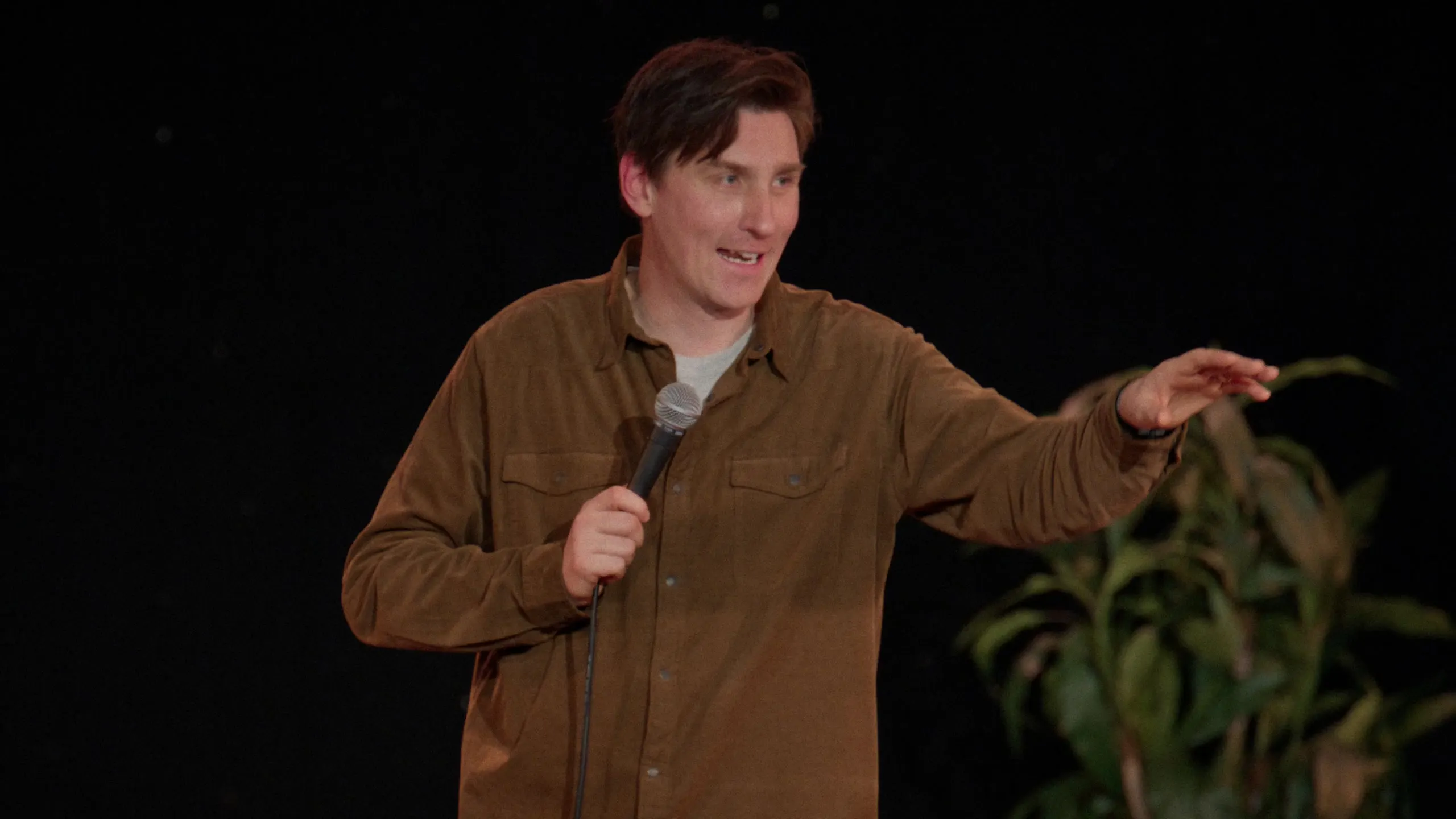 Sean McLoughlin mid-joke on stage, gesturing with the microphone in hand